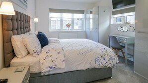 A bedroom with a bed, bedside lamps, a desk, a chair, a TV, and a window with a view. - Jasmine Cottage (Herne Bay)