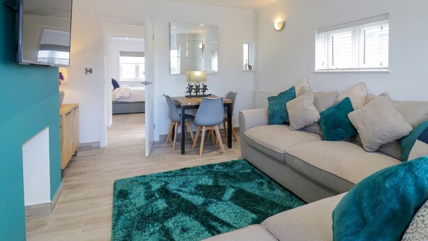 A modern living room with a sofa, a dining table with chairs, a TV mounted on the wall, and a large mirror. - Jasmine Cottage (Herne Bay)