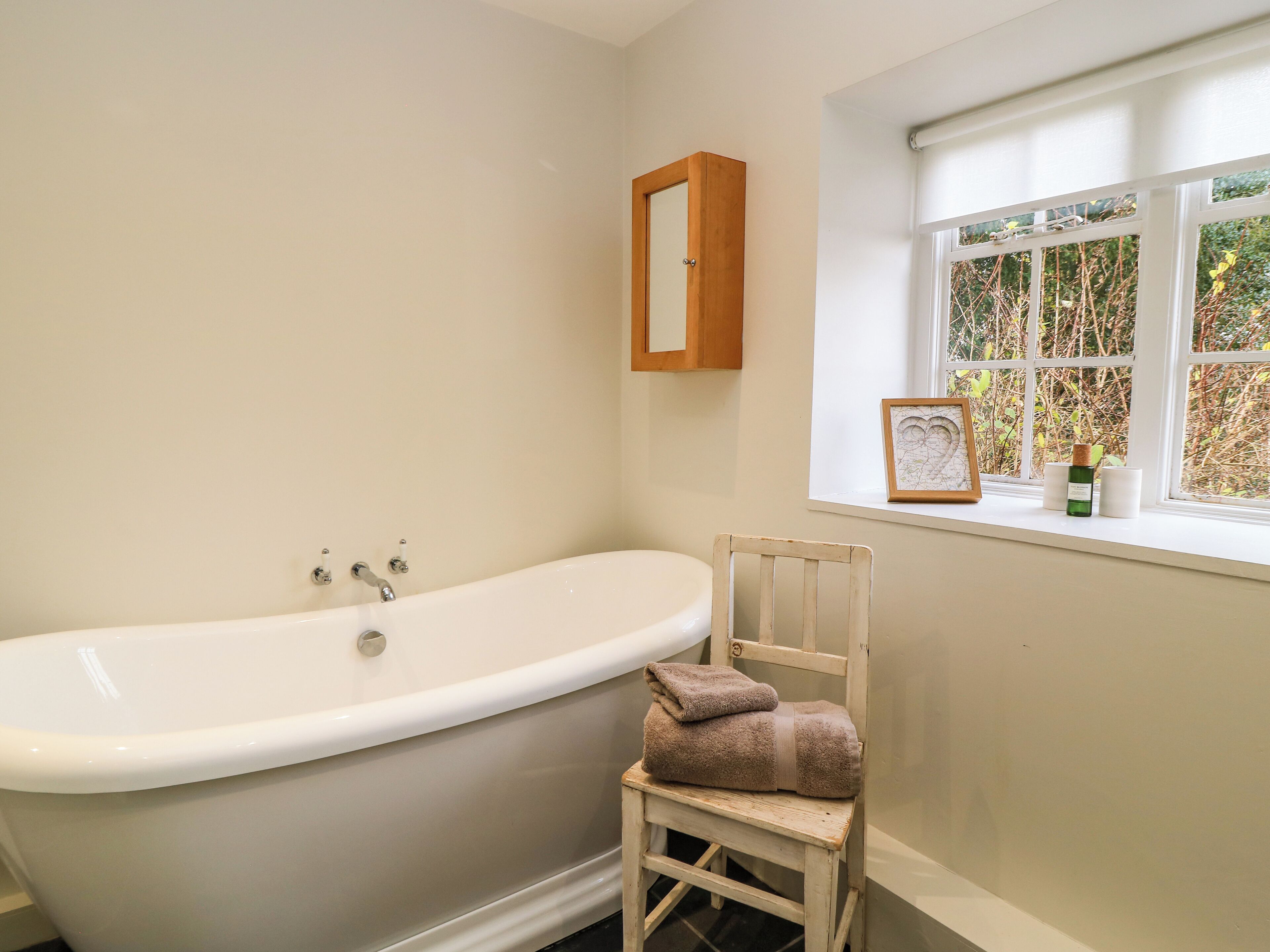 A bathroom with a freestanding bathtub, a wooden chair with a towel, a window with a view of greenery, and a wooden medicine cabinet.