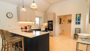 A modern kitchen with a large island, pendant lights, and a dining area with wicker chairs. - The Elm Coach House (Chipping Norton)