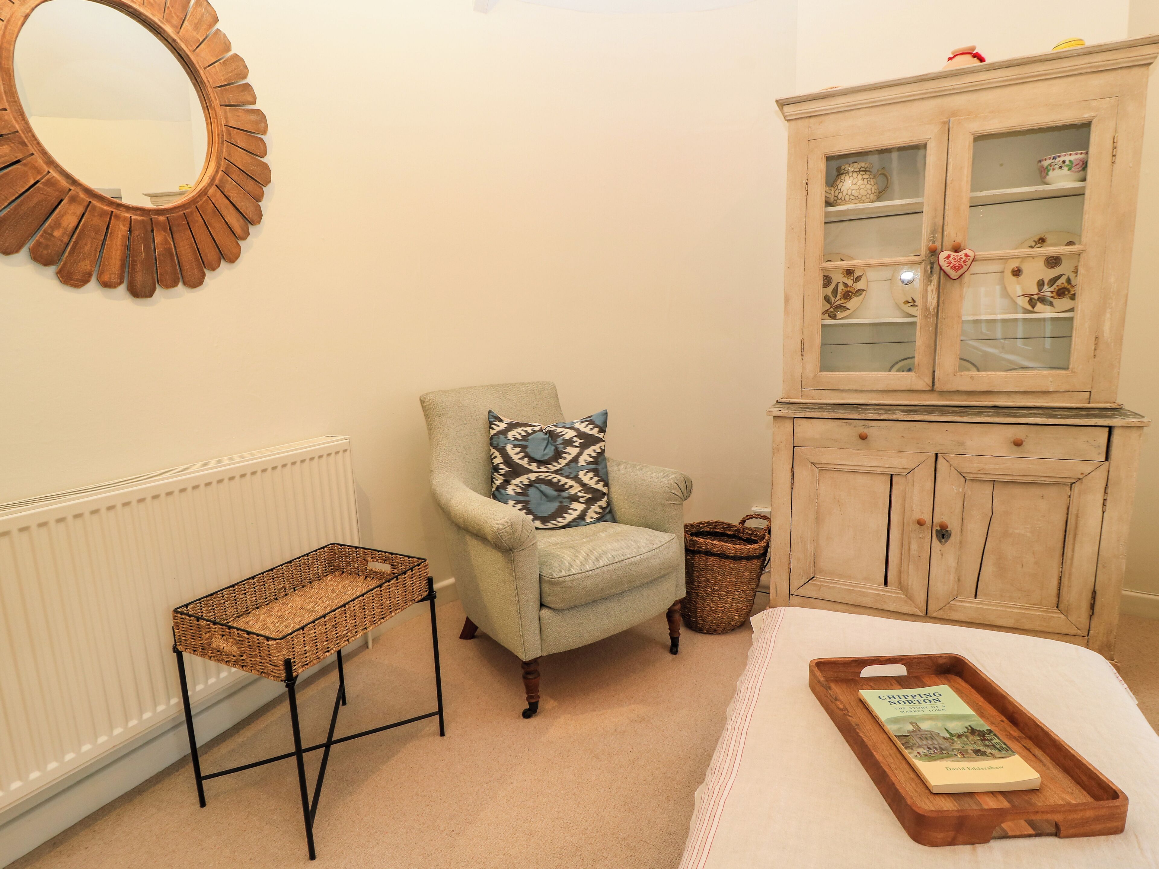 A cozy room with a radiator, a wicker side table, a cushioned armchair with a patterned pillow, a wooden cabinet with glass doors, and a wooden tray with a book on a bed.