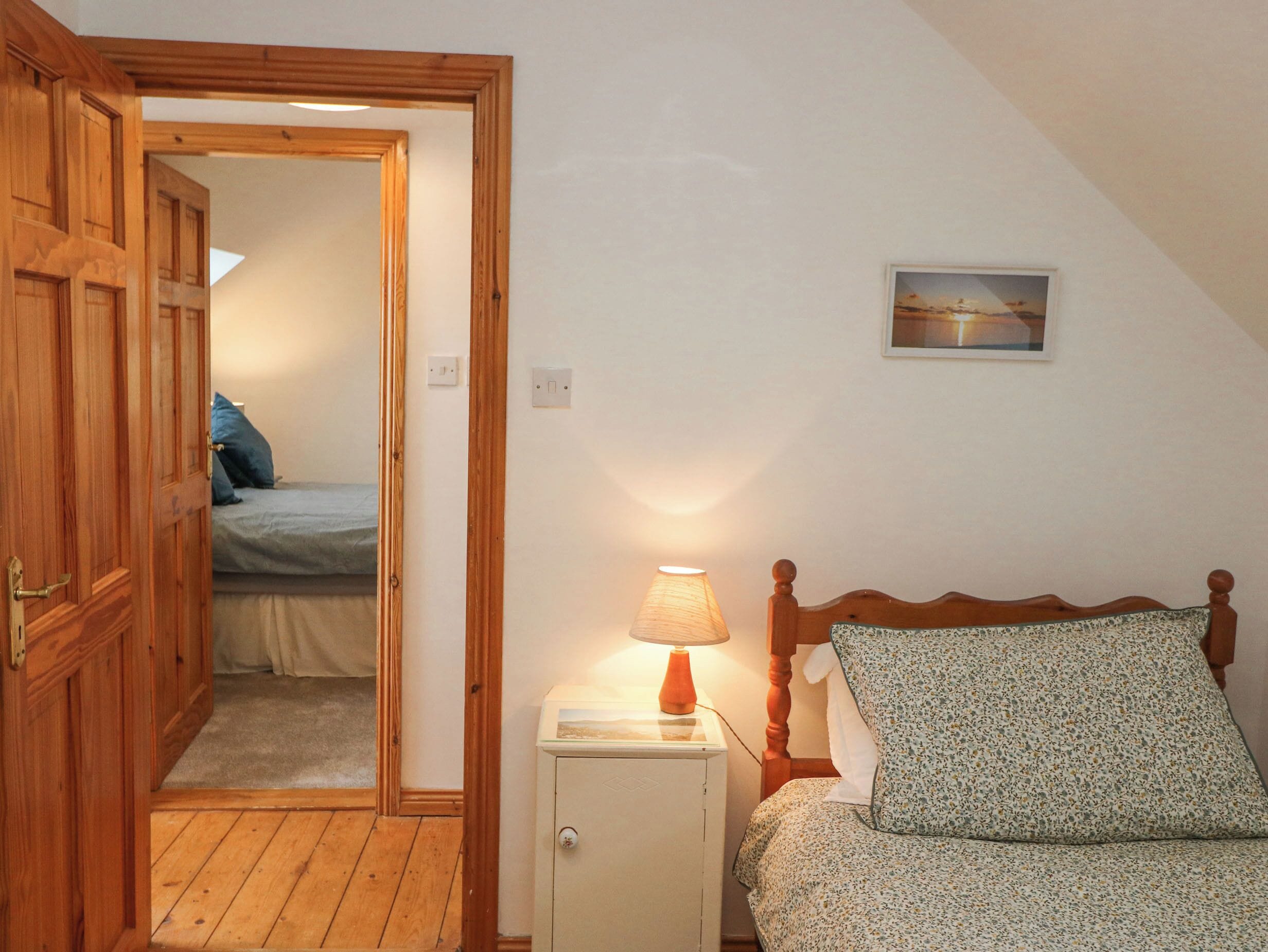 A bedroom with a wooden door, a bedside table with a lamp, a bed with a floral bedspread, and a painting on the wall.