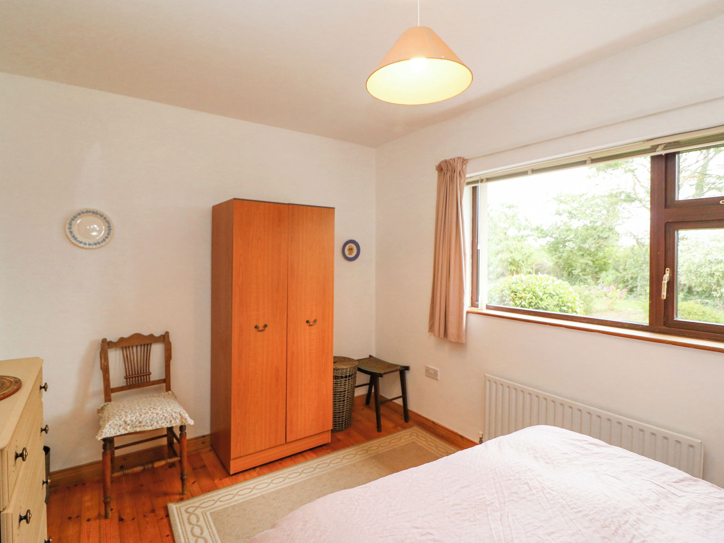 A bedroom with a wooden wardrobe, a chair, a small table, and a window with a view of greenery.