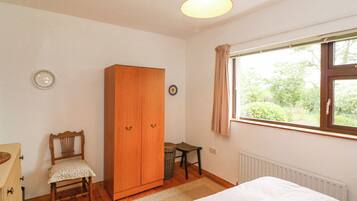 A bedroom with a wooden wardrobe, a chair, a small table, and a window with a view of greenery.