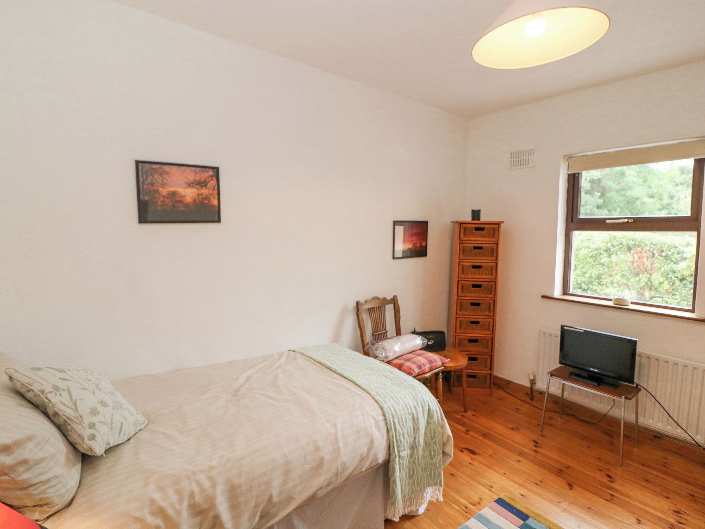 A bedroom with a bed, a chair, a TV on a stand, a window, and a wooden cabinet.