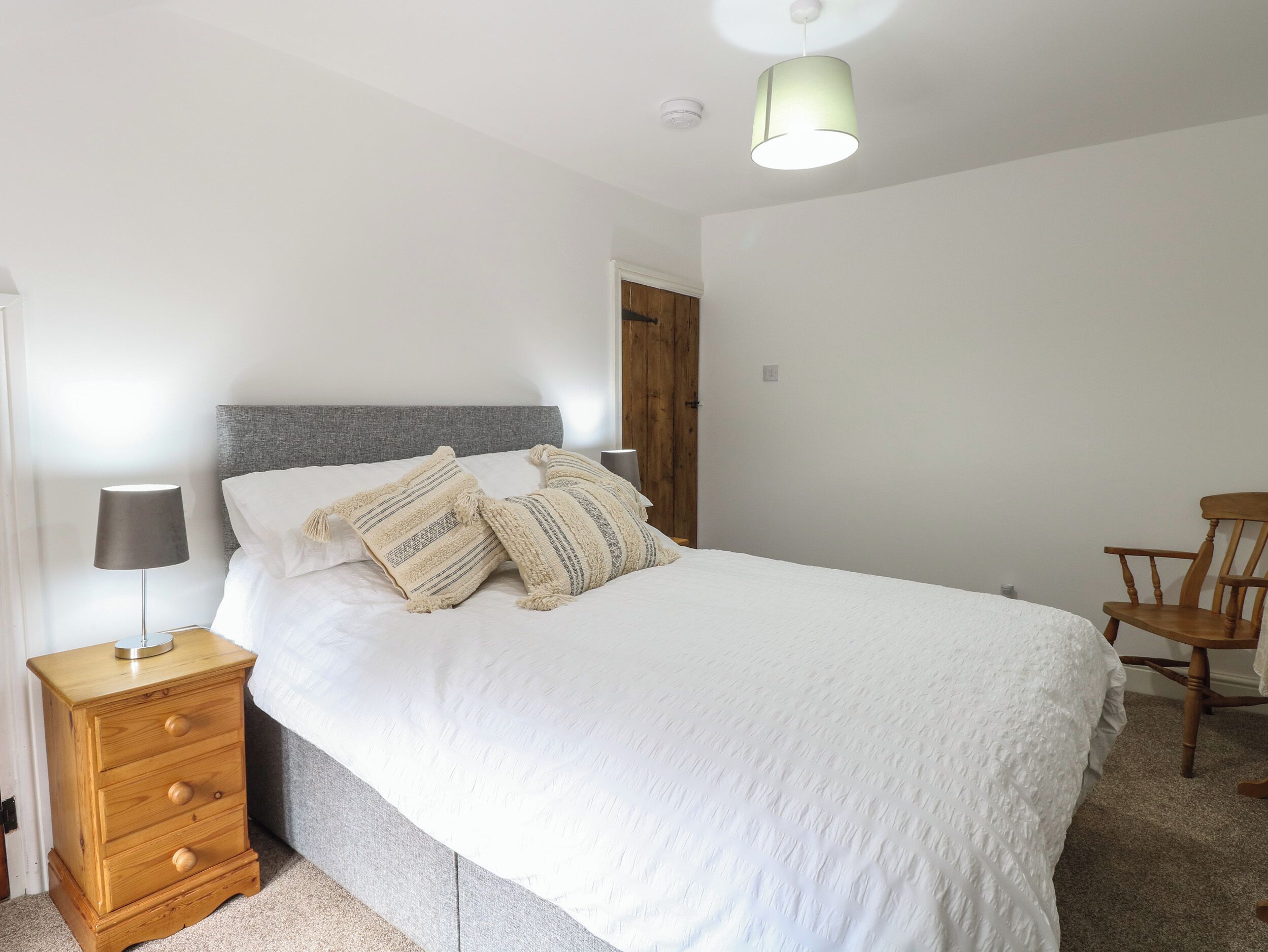 Una cama bien hecha con sábanas blancas y almohadones estampados, una mesita de noche de madera con una lámpara y una silla de madera en una habitación sencilla y luminosa.