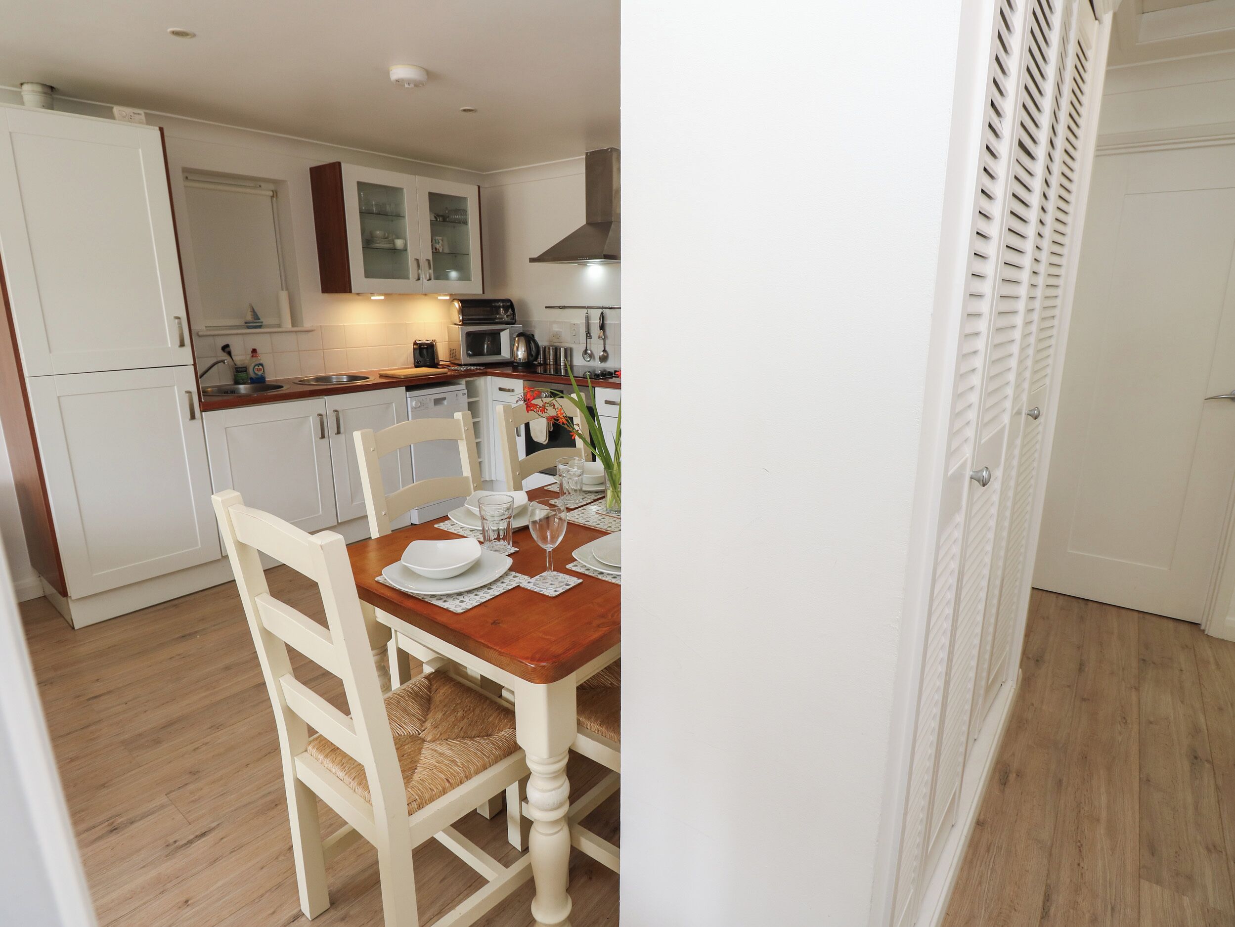Un coin repas avec une table en bois dressée pour quatre personnes, des chaises blanches et une cuisine en arrière-plan.