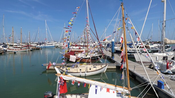 Un puerto deportivo con varias embarcaciones amarradas, con distintas banderas y un cielo despejado.