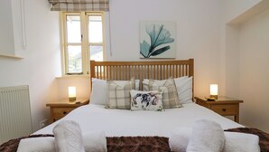 Ein ordentlich bezogenes Bett mit einer braunen Decke, zwei Nachttische mit Lampen, ein Fenster mit karierter Gardine und ein an der Wand hängendes Kunstwerk.