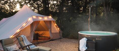 A tent with string lights, a hot tub, and lounge chairs in a forest setting.