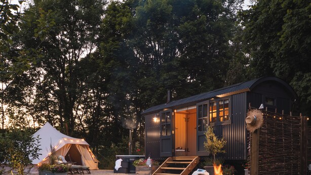 A rustic cabin with a large tent, a fire pit, and a wooden staircase leading to the entrance.