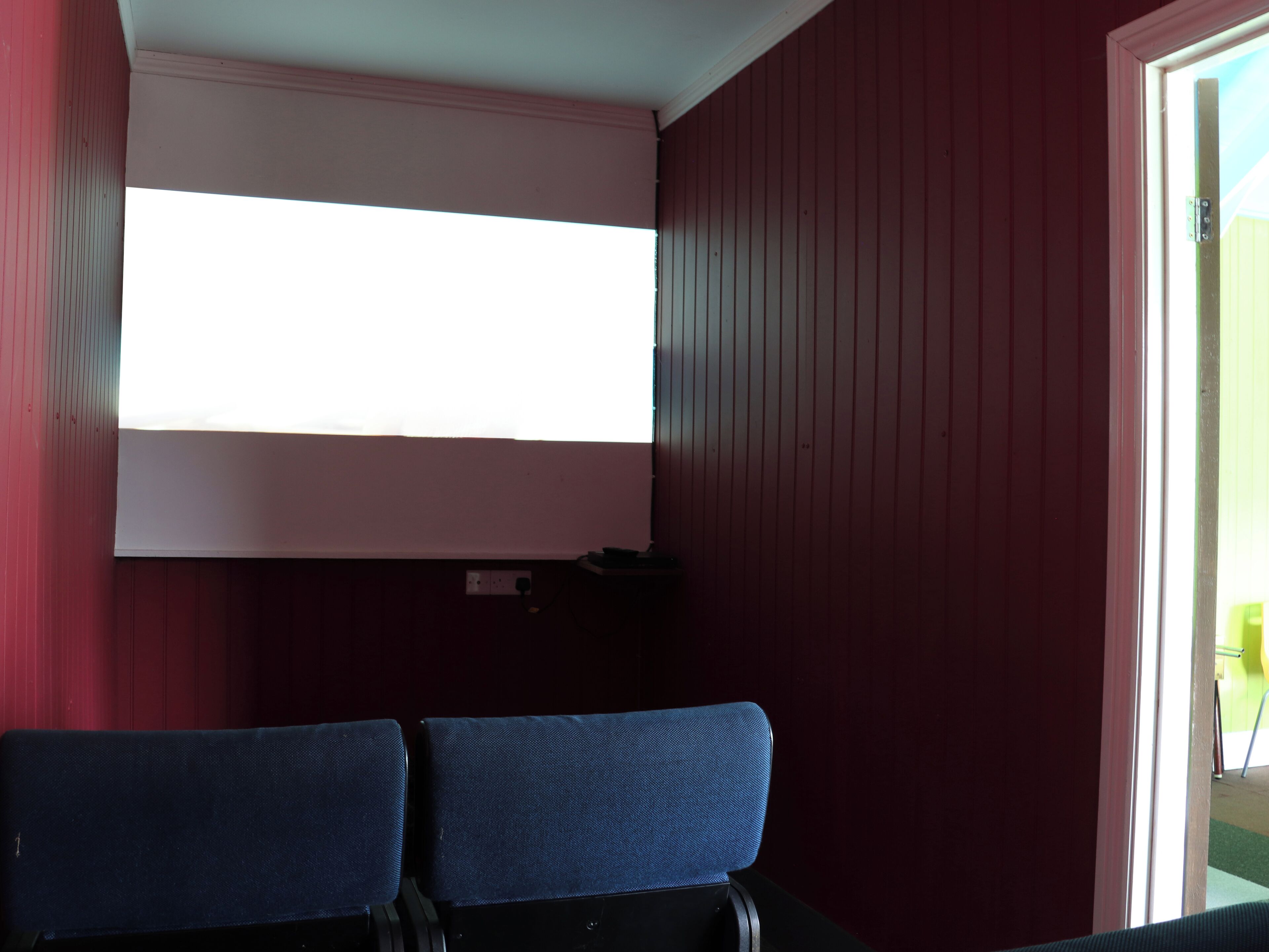 Una habitación con una pared roja, techo blanco y una ventana. Hay dos sillas azules y una mesa negra.