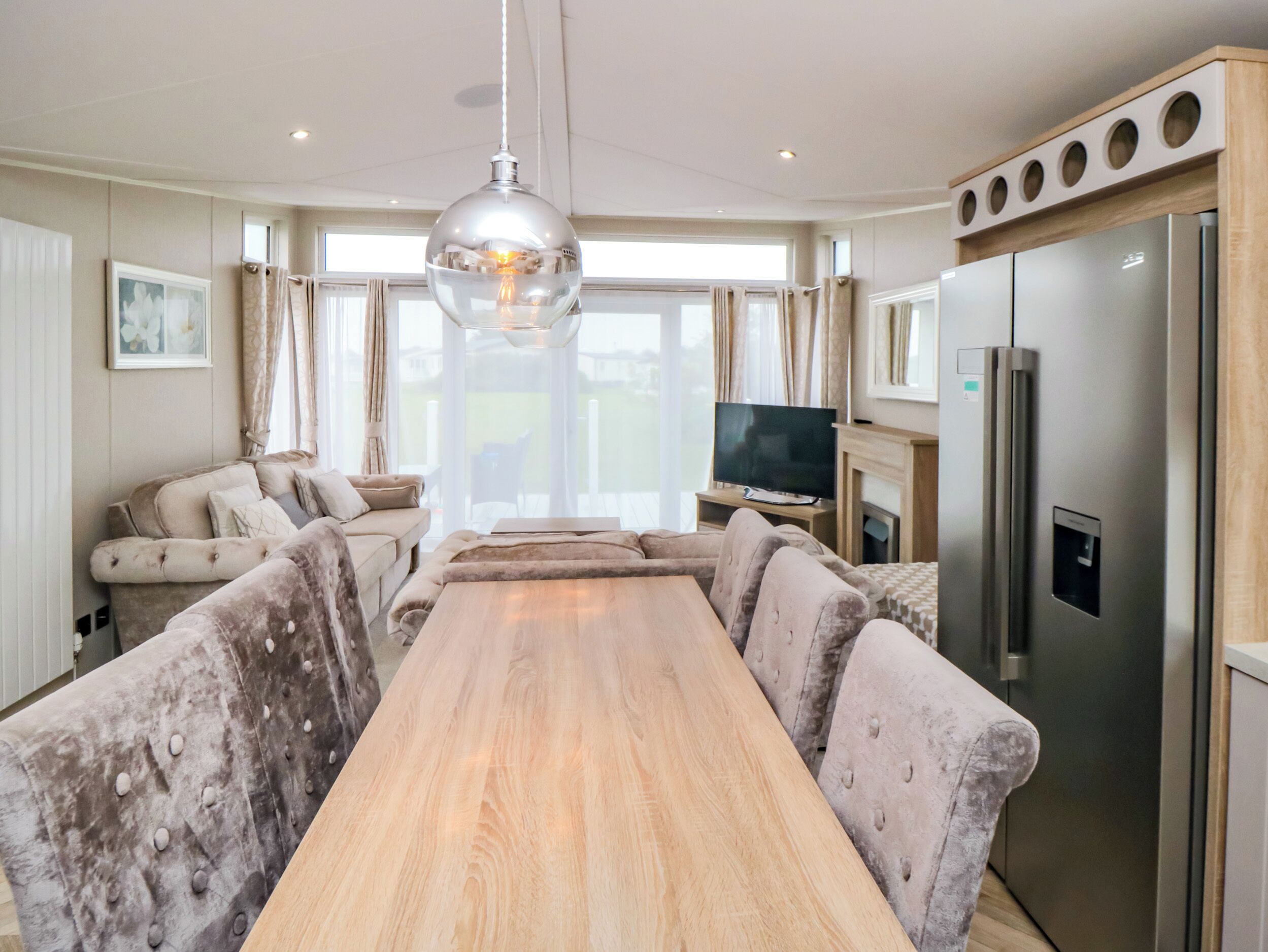 A modern living room with a long wooden table, plush seating, a large TV, and a built-in refrigerator.