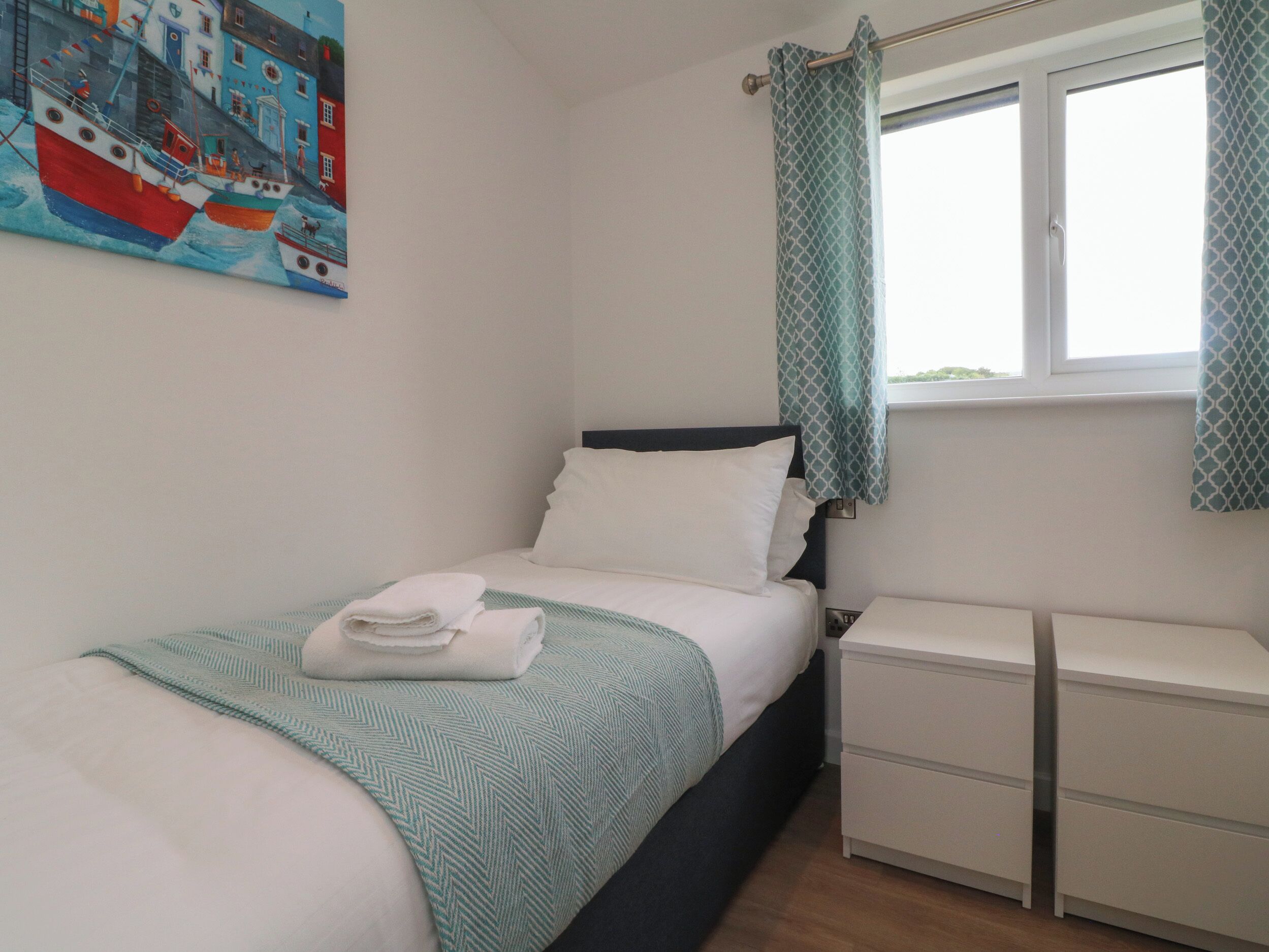 A neatly made bed with a white and teal striped blanket, a white pillow, and a folded white towel on the bed. There is a white bedside table with drawers and a window with a patterned curtain.