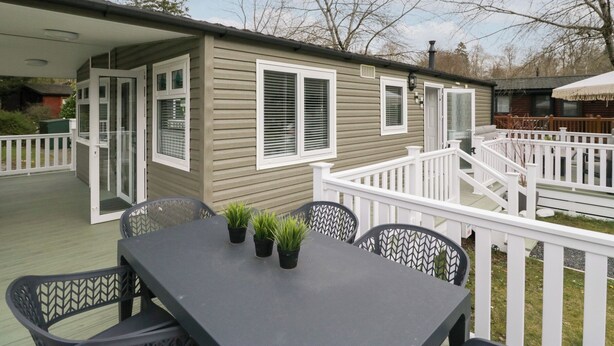 A covered patio area with a table and chairs, a small deck with railings, and a view of the surrounding trees and sky. - Cosy Corner (Windermere)