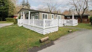 A two-story mobile home with a porch and a small deck, surrounded by a grassy yard and trees. - Cosy Corner (Windermere)