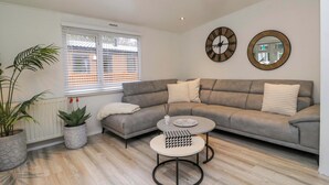 A modern living room with a grey sectional sofa, a round coffee table, and decorative pillows. There are two wall clocks and a mirror. - Cosy Corner (Windermere)