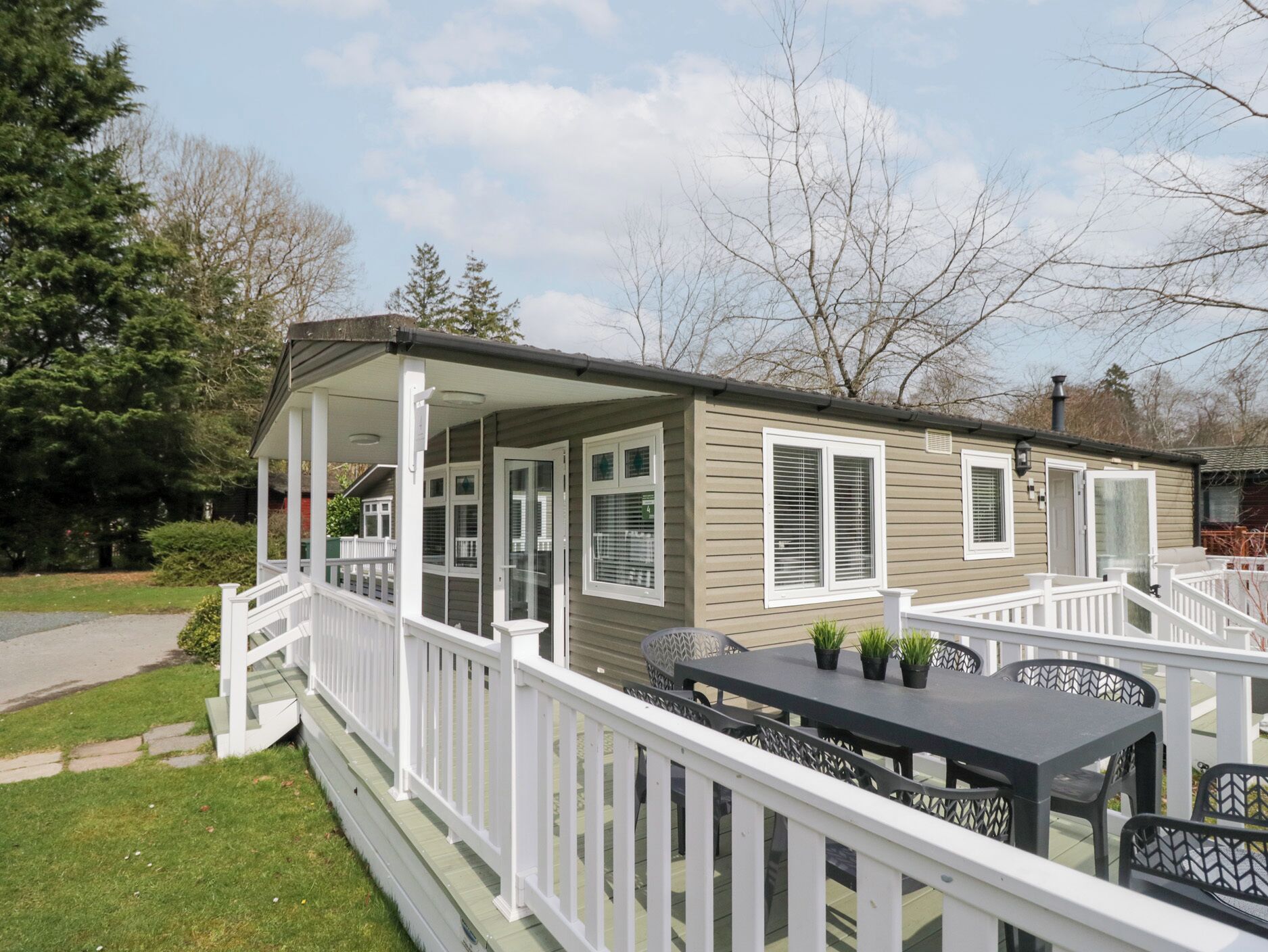 A single-story mobile home with a covered porch, white railings, and a patio area with a table and chairs.