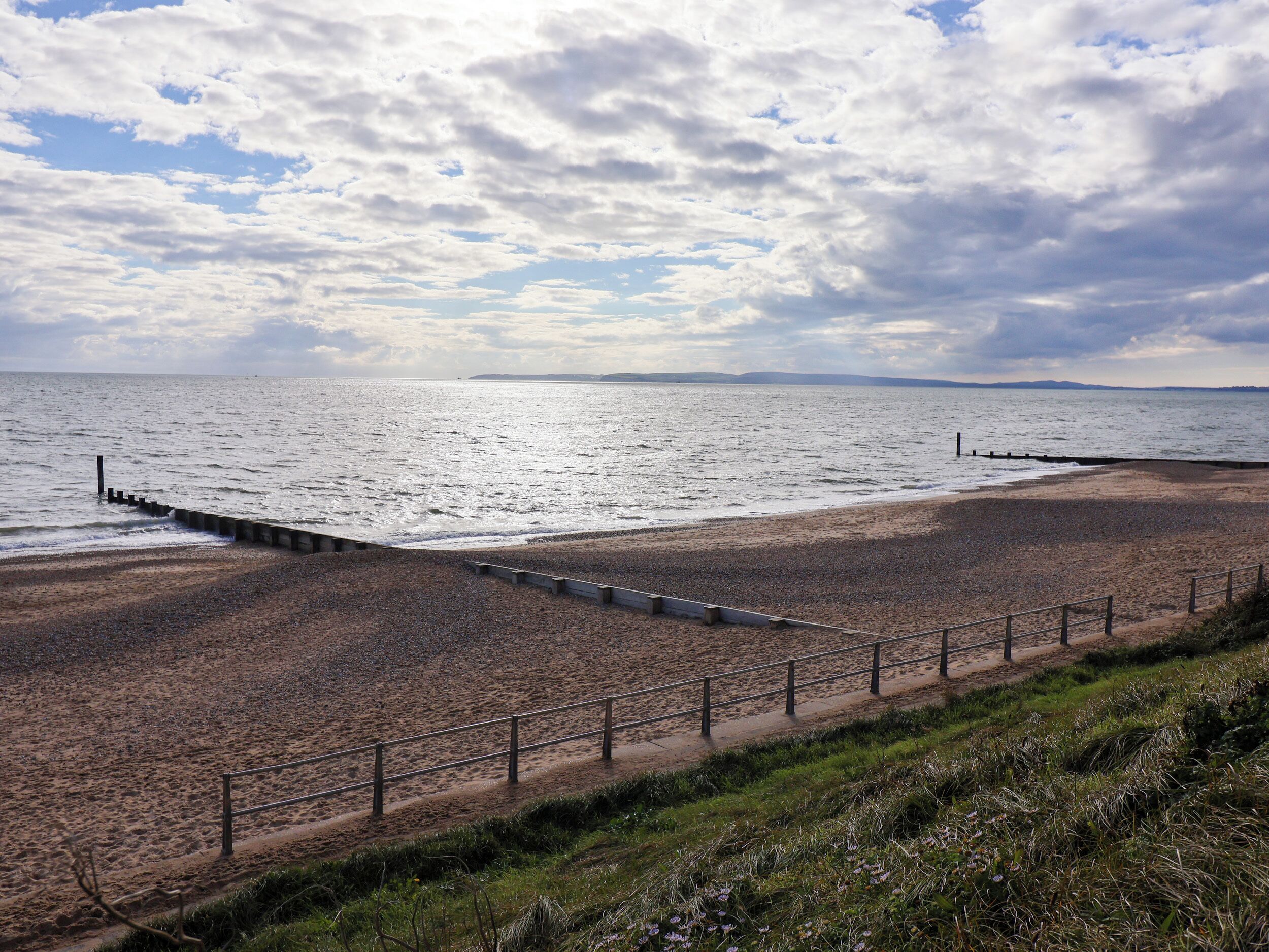 Une plage avec une promenade en bois menant à l’eau, une clôture le long du bord et une zone herbeuse au premier plan.