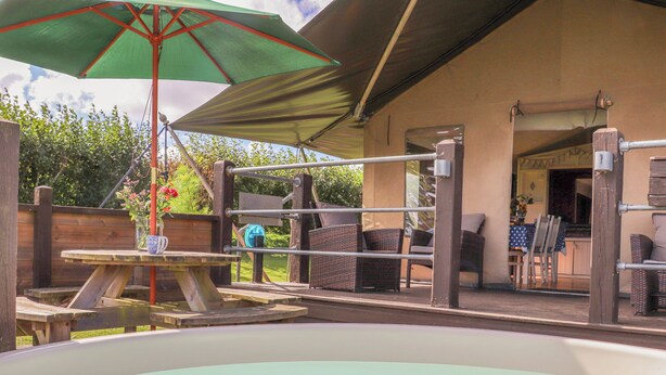 An outdoor hot tub area with a table and chairs under a shaded umbrella, surrounded by wicker furniture and a wooden fence. - Bluebell (Penzance)