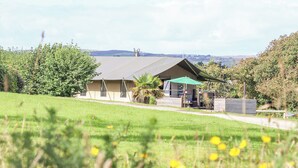 A single-story building with a sloped roof, a patio area with a table and chairs, and a view of the countryside. - Bluebell (Penzance)