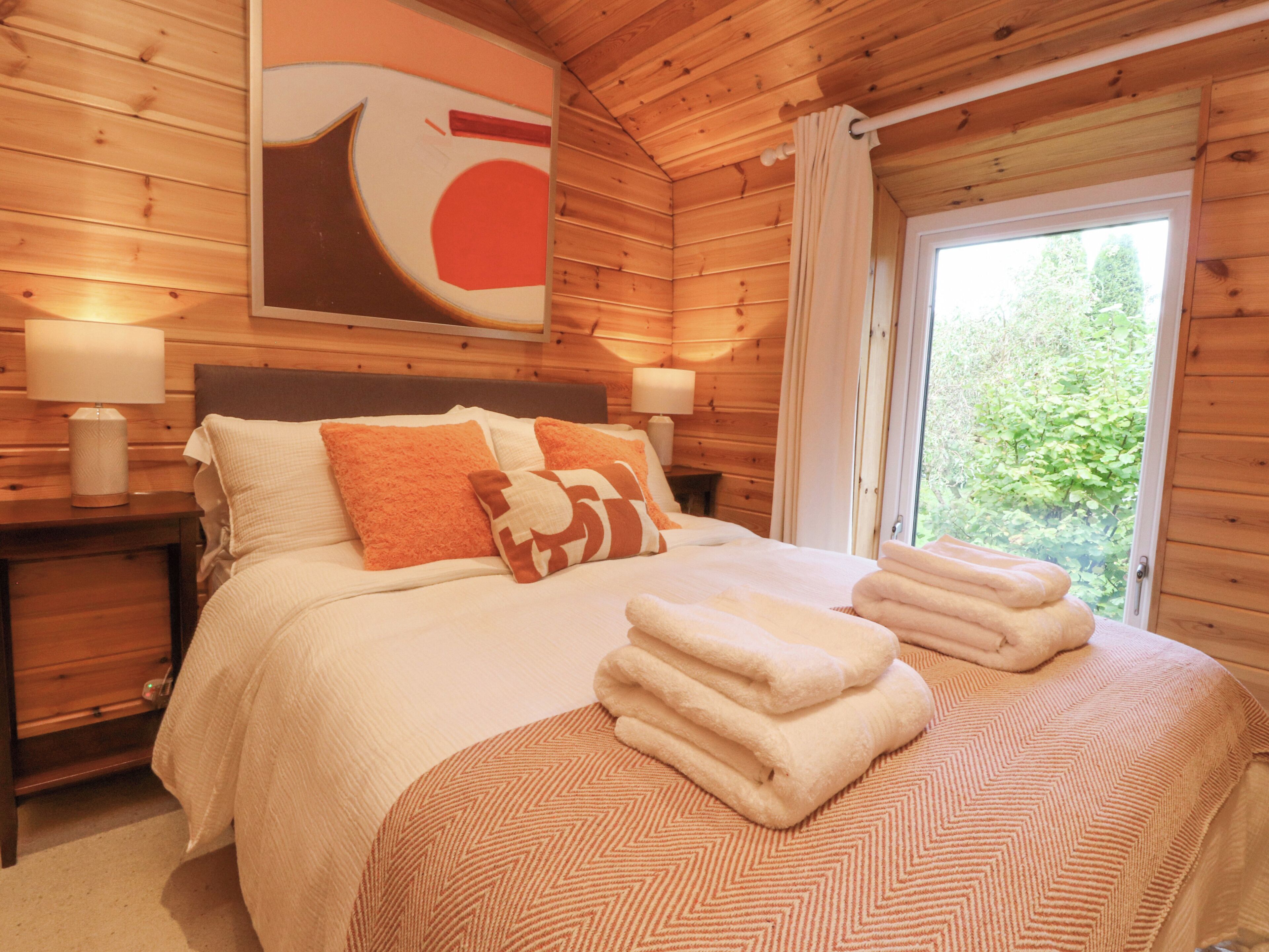 A wooden cabin bedroom with a bed, pillows, a nightstand, and a window with curtains.