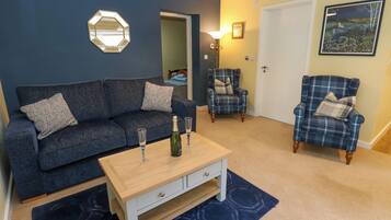 Uma sala de estar com um sofá azul escuro, uma mesa de centro de madeira, duas poltronas e um espelho hexagonal na parede.