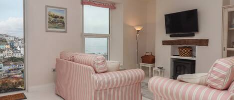 A living room with striped sofas, a television, a fireplace, and a view of a coastal town.