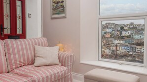 A cozy room with a striped sofa, a small ottoman, a framed painting, and a window with a view of a colorful town. - The White House (Brixham)