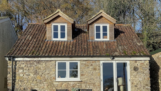 Una casa in pietra con tetto a tegole, due finestrelle a mansarda, un portone d'ingresso e un piccolo patio con tavolo e sedie.