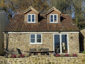 Una casa de piedra con techo de tejas, dos ventanas salientes, una puerta principal y un pequeño patio con una mesa y sillas.