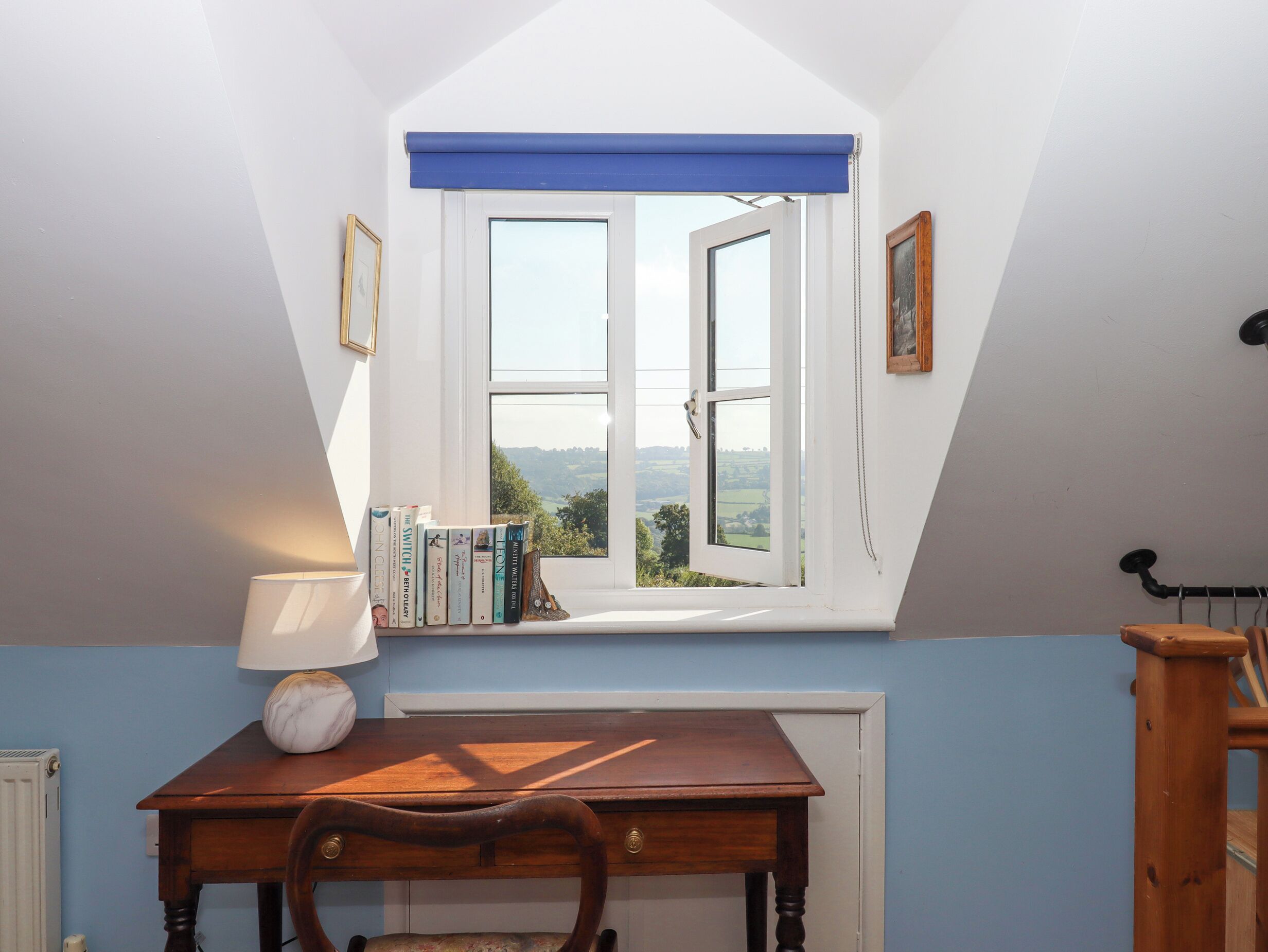 Une pièce avec un plafond en pente, une fenêtre avec un store bleu, une table en bois avec une lampe et une bibliothèque remplie de livres.