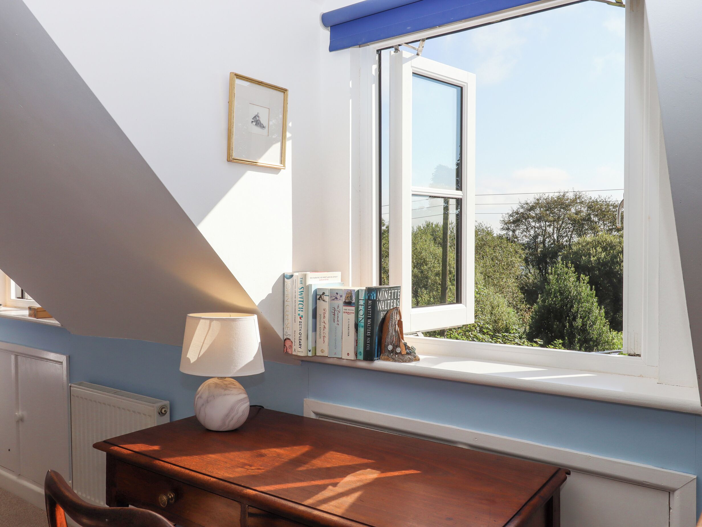 A room with a sloped ceiling, a window with a view of greenery, a table with a lamp, and books on a ledge.