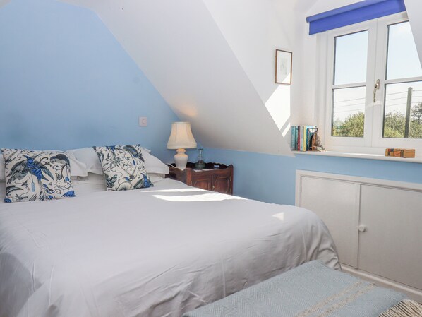 A bedroom with a sloped ceiling, a bed with white bedding, a nightstand with a lamp, a window with books on the sill, and a radiator. - The Wheelhouse (Honiton)