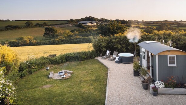 Een rustieke blokhut met een jacuzzi, omringd door een grindpad, weelderige begroeiing en uitzicht op glooiende heuvels.