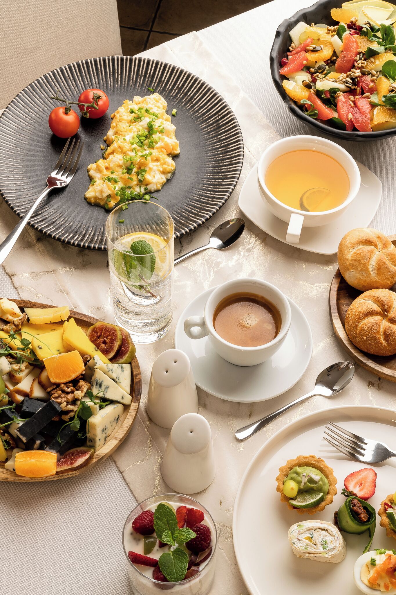 Mesa posta para o pequeno-almoço com ovos mexidos, salada de frutas, pastelaria e uma chávena de café.
