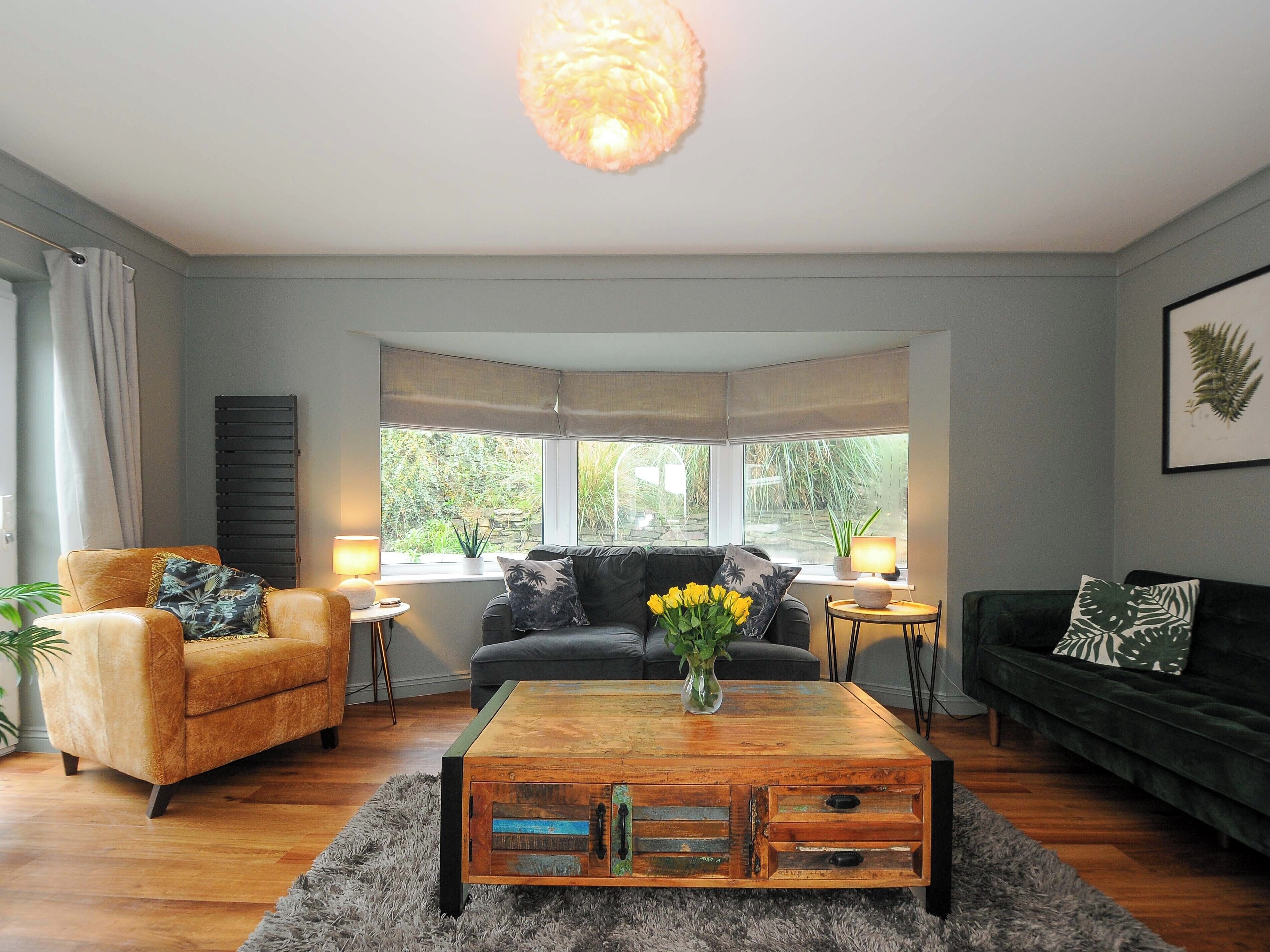 A living room with a sofa, armchair, and coffee table. There are windows with curtains, a plant, and a lamp.