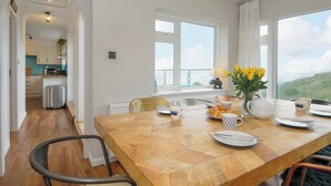 Zona pranzo con tavolo in legno apparecchiato per quattro persone, un vaso con fiori gialli e vista sull'esterno attraverso ampie finestre.