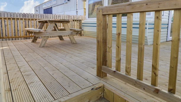 A wooden deck with picnic tables, a metal fence, and a building with a white exterior and windows. - Meric Stone Holiday Lets Caravan 2 (Limavady)