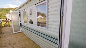 A mobile home with a wooden deck, a sliding door, and a window with a view of a house and a hill. - Meric Stone Holiday Lets Caravan 2 (Limavady)