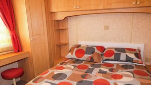 A bedroom with a bed, a red chair, wooden cabinets, and a window with red curtains. - Meric Stone Holiday Lets Caravan 2 (Limavady)
