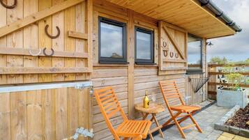 A wooden cabin with two windows, a door, and a covered patio area with two orange chairs and a small table.