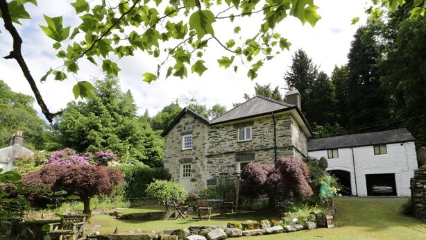 A stone house with a stone exterior, a small garden with a wooden bench, and a car parked in the driveway. - Beaver Grove Cottage (Betws-Y-Coed)