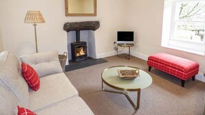 A living room with a sofa, a red bench, a small table with a tray, a fireplace, a television, and a window with a view. - Beaver Grove Cottage (Betws-Y-Coed)