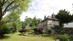 A stone house with a stone chimney, surrounded by a well-maintained garden with various shrubs and a lawn. - Beaver Grove Cottage (Betws-Y-Coed)
