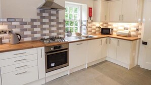 A modern kitchen with white cabinets, a gas stove, an oven, a microwave, and a window with a view of greenery. - Beaver Grove Cottage (Betws-Y-Coed)