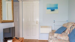 A room with a wooden table, a television, a white door, a radiator, and a floral sofa. - High Cove (Wadebridge)