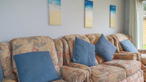 A row of floral-patterned sofas with blue cushions, a light blue wall, and three vertical paintings depicting beach scenes. - High Cove (Wadebridge)