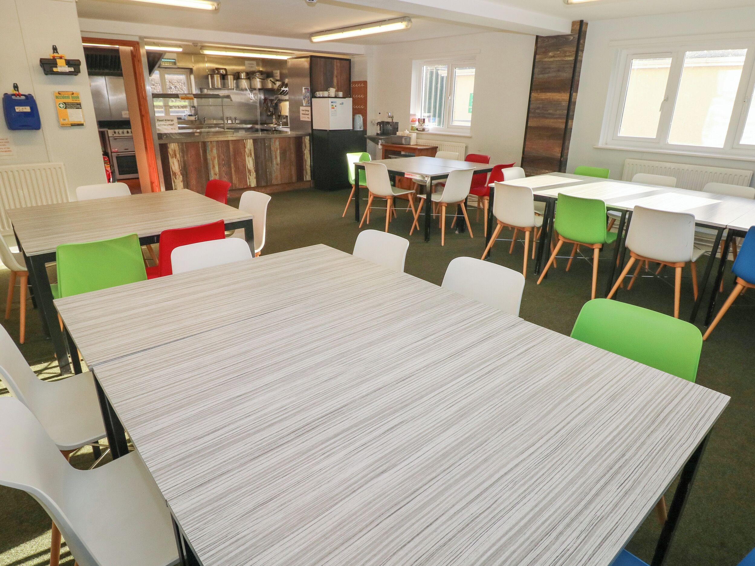 A dining area with tables and chairs, a kitchen in the background, and windows with blinds.