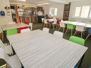 Un espace repas avec des tables et des chaises, une cuisine à l’arrière-plan et des fenêtres équipées de stores.